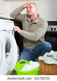 Man Is Holding His Head Sitting Near Washing Machine Thinking Of How To Use It. Guy Doing Laundry