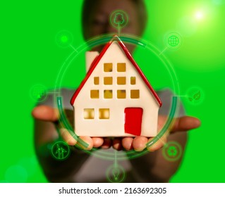 Man Holding House In His Hands With Alternative Green Renewable Power Sources, Solar Energy, Bio Fuel On Blurred Background, Eco-friendly Housing Concept.