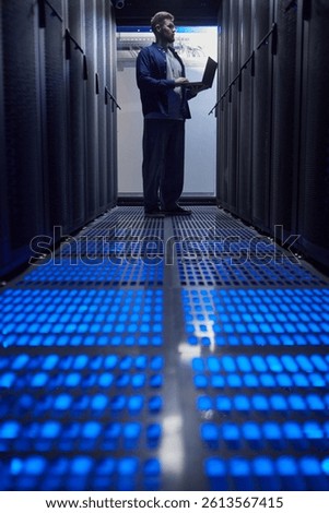 Man holding laptop in a data server room, surrounded by tall server racks, with blue illuminated floor panels creating a futuristic atmosphere