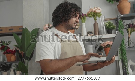 Man holding laptop and typing while arranging a bouquet in studio flower shop, wearing apron and checking orders; focused small business care.