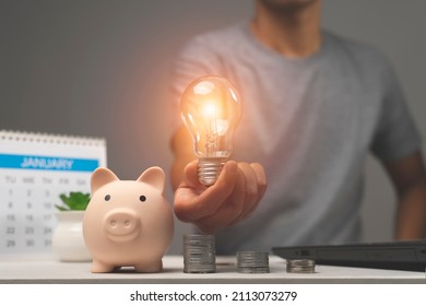 Man Holding Lightbulb On Piggy Bank And Coins Stacking On Desk. Saving Money Concept