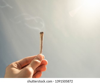 Man holding a lit joint of marijuana with a soft sky and shiny sun in the background. Cigarette with smoke, right and top copy space.