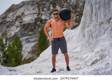 Man Holding Medicine Ball in Quarry. A man wearing athletic shorts and sunglasses stands in a quarry, holding a medicine ball on his shoulder. - Powered by Shutterstock