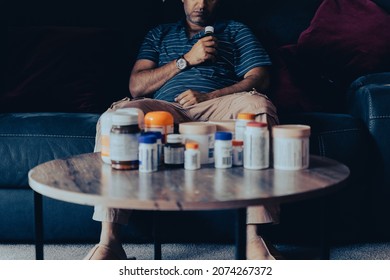 Man Holding Medicine Bottle Sitting On Sofa. Sad Person Sitting On Couch With Several Medicine Bottles. Concept Of Sickness. 