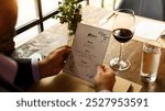 Man holding a menu at a restaurant table with a glass of red wine and water. Man reading menu with wine at the restaurant. Menu read by a man at restaurant