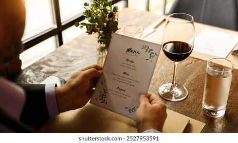 Man holding a menu at a restaurant table with a glass of red wine and water. Man reading menu with wine at the restaurant. Menu read by a man at restaurant's table with glass of wine and water. - Powered by Shutterstock