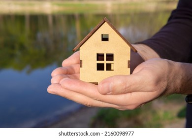 Man holding a miniature wooden house model with both hands against a lake background. Home insurance and coverage concept. - Shutterstock ID 2160098977