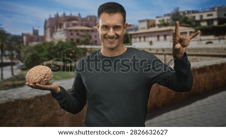 Man holding a model brain in left hand while making rock horns with right hand on a street beside historic building; playful curiosity.