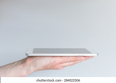 Man Holding Modern Silver And White Tablet Or Smart Phone In One Hand, Viewed From The Side With Real Estate Above The Display For Additional Information. Grey Background.