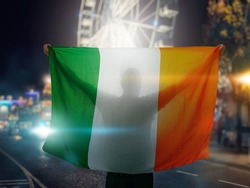 Man Holding National Flag Of Ireland In His Hands. Christmas Themed Background Out Of Focus. Celebrating Festive Season. Male Fan With Irish Flag. Protest Or Support Concept.