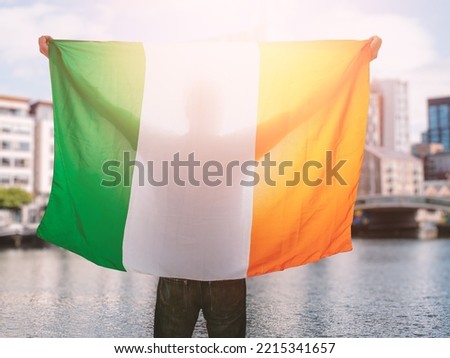 Man holding National flag of Ireland in focus. Dublin city out of focus in the background. Support or protest concept. Sun flare. Male with Irish flag.