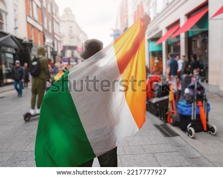 Man holding National flag of Ireland in focus. Grafton street of Dublin city out of focus in the background. Support or protest concept. Sun flare. Male with Irish flag.