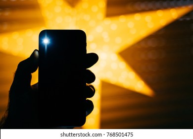 Man Holding The New Phone With Yellow Star Bokeh Background Featuring A Flare Flash Light From The Rear Flash And Camera - Holiday Concept 