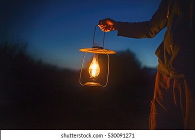 Man Holding The Old Lamp With A Candle Outdoors. Hand Holds A Large Lamp In The Dark. Ancient Lantern With A Candle Illuminates The Way On A Night