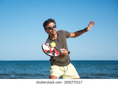 Man Holding A Paddle Tennis Racket Hitting The Ball On A Blue Background. Young Sportsman Playing Tennis On The Beach. Paddle Player