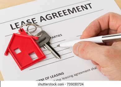 Man Holding Pen Over House Door Key With Red Keychain Pendant And Lease Agreement Form On Wooden Desk - House Or Apartment Rental Concept