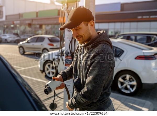 Man Holding Power\
Charging Cable For Electric Car In Outdoor Car Park. And he s going\
to connect the car to the charging station in the parking lot near\
the shopping center