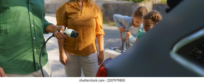 Man Holding Power Supply Cable, While His Family Waiting For Car Charging At Electric Vehicle Charging Station, Closeup