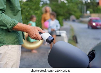 Man Holding Power Supply Cable, While His Family Waiting For Car Charging At Electric Vehicle Charging Station, Closeup