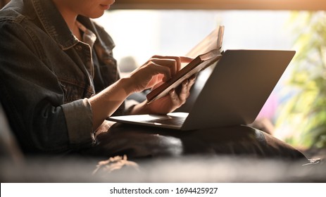 Man Holding And Reading A Book While Sitting In Front A Computer Laptop At The Leather Couch Over Comfortable Living Room As Background.