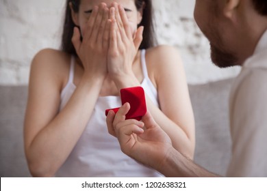 Man holding red box with marriage engagement ring, preparation make proposal to loving woman, girlfriend sitting on couch covered face in hands, boyfriend standing on knee, marry me concept close up