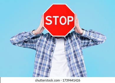 Man Is Holding Red Prohibition Road Sign Stop In Front Of Face On Blue Background. Guy Is Warning About Danger, Risk. Prevention Symbol, Attention Icon That Shows Traffic Ban. Caution, Alert Concept.