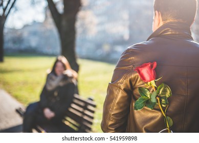 Man Holding A Red Rose Behind Back, Woman Siting On The Park Bench In The Background