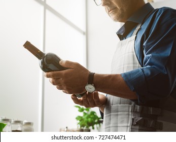 Man Holding A Red Wine Bottle And Reading The Label, He Is Cooking In The Kitchen At Home