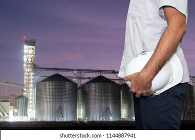 A man holding safety helmet. in the background set of storage tanks raw material agricultural crops feed mills at twilight. Safety at work