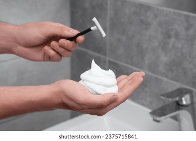Man holding shaving foam and razor in bathroom, closeup