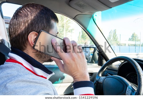 A man\
holding a silver mobile phone near his ear and talking on it while\
driving. Danger of using the phone in a\
car