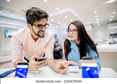 Man holding smart phone and pointing on other phone which  is in woman`s hands. Tech store interior.