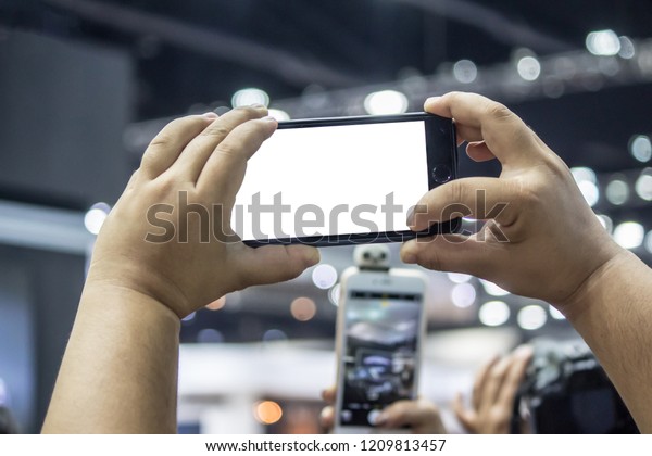 man  holding smartphone to take photo with her hands\
in a exhibition hall with group of people. Reporters waiting for\
press conference live