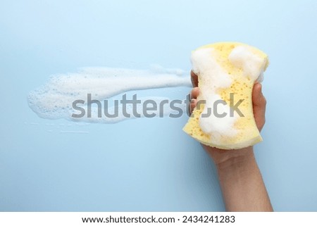 Man holding sponge with foam on light blue background, top view
