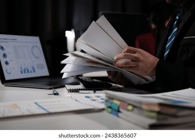 A man is holding a stack of papers and looking at a laptop. The scene suggests that he is working on a project or analyzing data