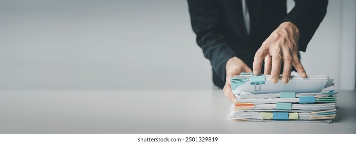 A man is holding a stack of papers on a table. The papers are piled high and the man is reaching for one. Concept of urgency and importance