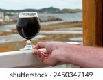 A man holding a stout beer in a clear tulip shaped glass on the railing of a patio. There