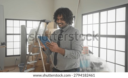 Man holding tablet tapping screen inside building amid packed boxes and ladder during new home walkthrough; optimism future plans.