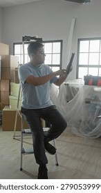 Man holding tablet while seated on a step ladder amid moving boxes, toolbox and plastic covered furniture in building; reflective.