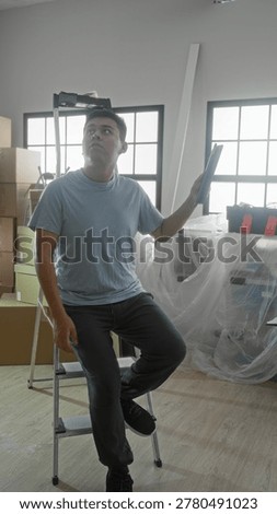 Man holding tablet while seated on a step ladder amid moving boxes, toolbox and plastic covered furniture in building; reflective.