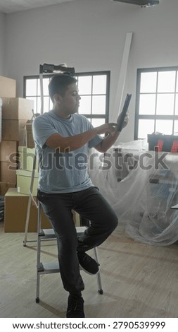 Man holding tablet while seated on a step ladder amid moving boxes, toolbox and plastic covered furniture in building; reflective.