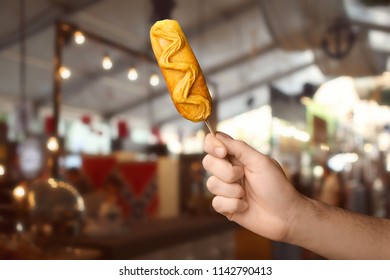 Man Holding Tasty Corn Dog With Mustard In Cafe