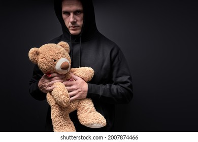 Man Holding Teddy In Hands, Toy Is Simulating Child Abuse, Rape And Kidnapping. Adult Man In Casual Black Clothes Having Frightening Look. Isolated On Black Studio Background. Copy Space