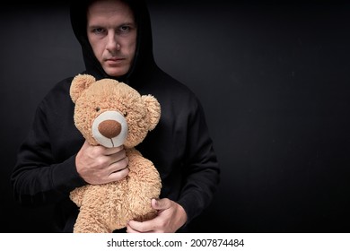 Man Holding Teddy In Hands, Toy Is Simulating Child Abuse, Rape And Kidnapping. Adult Man In Casual Black Clothes Isolated On Black Studio Background. Copy Space