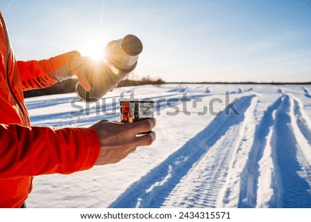 Man holding thermos with cup of tea, winter landscape, setting sun shining reflections on the camera, trekking in the forest, winter hike in the snow. High quality photo