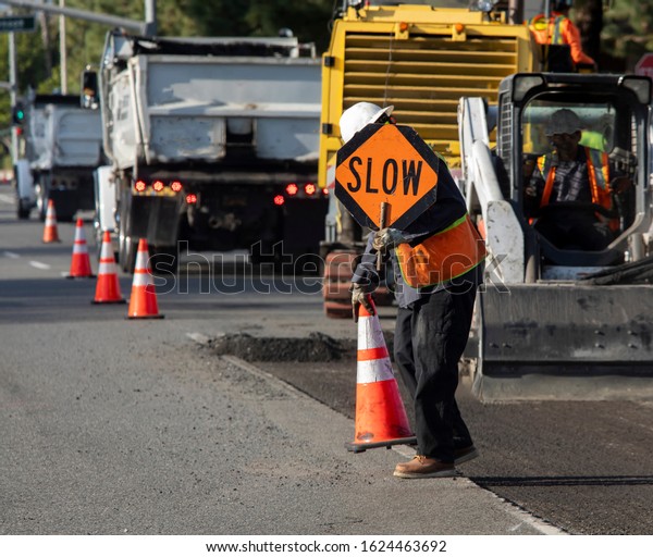 Man holding traffic sign that says SLOW at a\
roadway construction site. Bright orange traffic cones and trucks\
and cars are visible