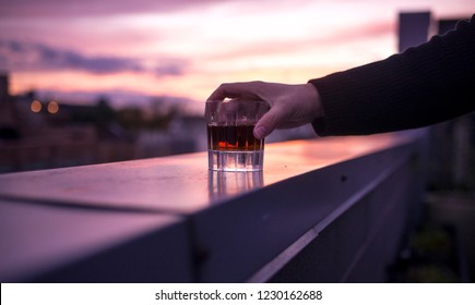 A Man Holding A Tumbler Of Premium Whkskey On A Rooftop Bar During A Colorful Sunset.