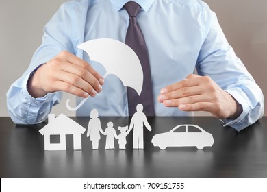 Man Holding Umbrella Over Paper Silhouette Of Family, House And Car On Table. Insurance Concept