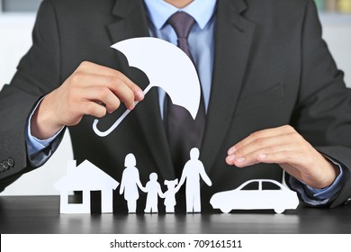 Man Holding Umbrella Over Paper Silhouette Of Family, House And Car On Table. Insurance Concept