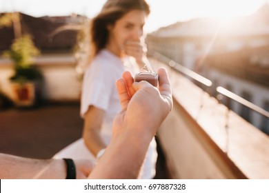 Man holding wedding ring in front of astonished happy girl covering mouth with hand. Romantic photo of charming woman standing on roof early in evening on date with boyfriend in anniversary. - Powered by Shutterstock